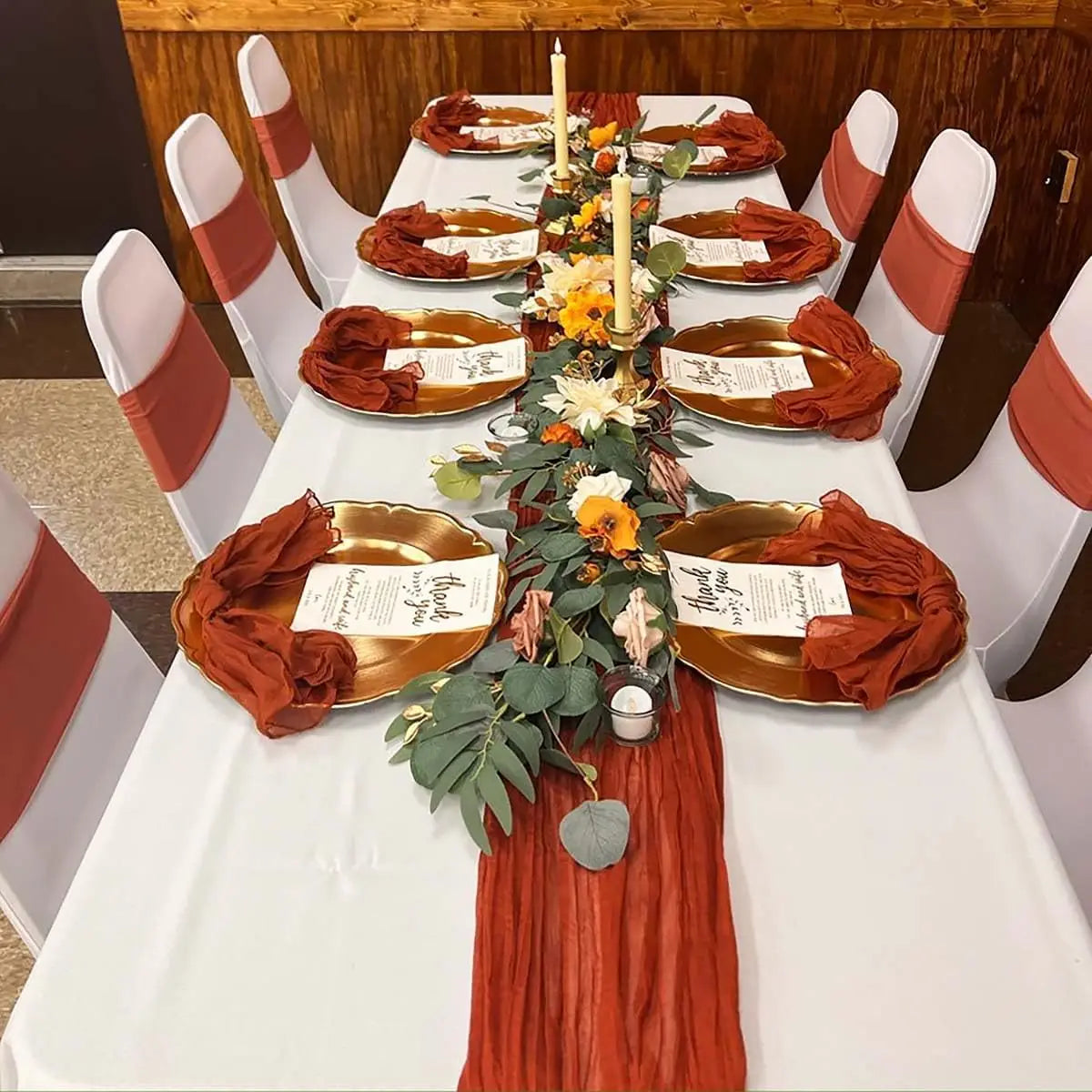 A long white table is set with terracotta-colored plates, candles, and greenery for an event.