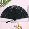 Black folding hand fan with white bamboo design and tassel shown against a pink background.