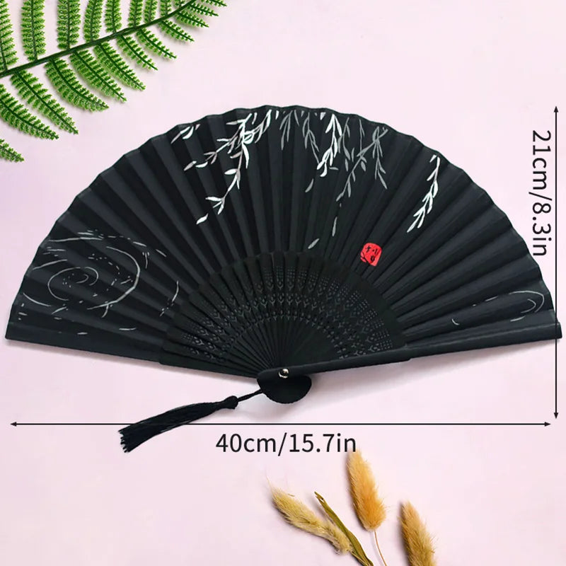 Black folding hand fan with white bamboo design and tassel shown against a pink background.