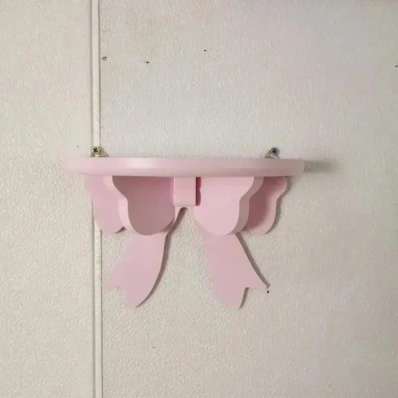 A pink decorative bow-shaped wooden wall shelf mounted on a white wall.