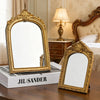 A vintage gold-framed arched vanity mirror resting on a wooden table.