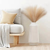 A tall white vase holds a fluffy beige pampas grass floral decor arrangement next to a light gray sofa.