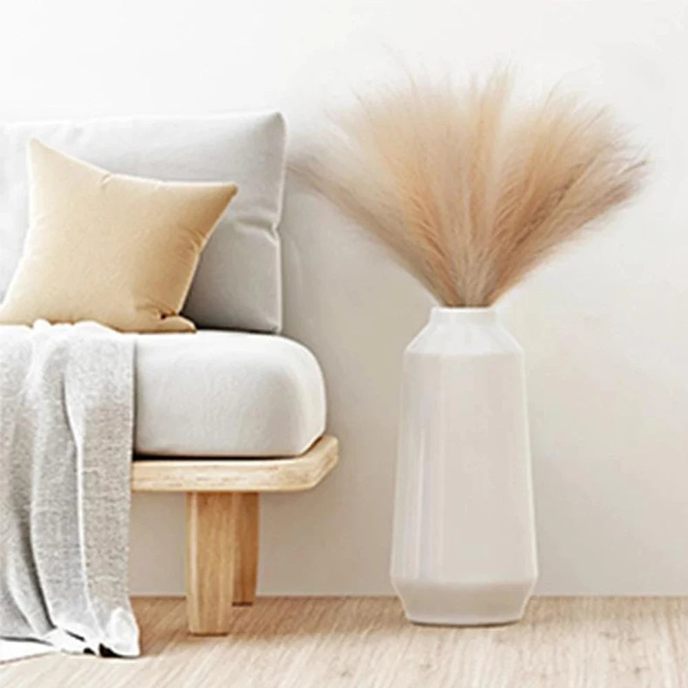 A tall white vase holds a fluffy beige pampas grass floral decor arrangement next to a light gray sofa.