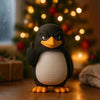A black and white penguin desk statue with a raised flipper sitting on a wooden surface in front of a blurred Christmas tree.