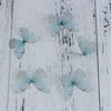 Four light blue organza butterfly embellishments scattered on a white wooden surface.
