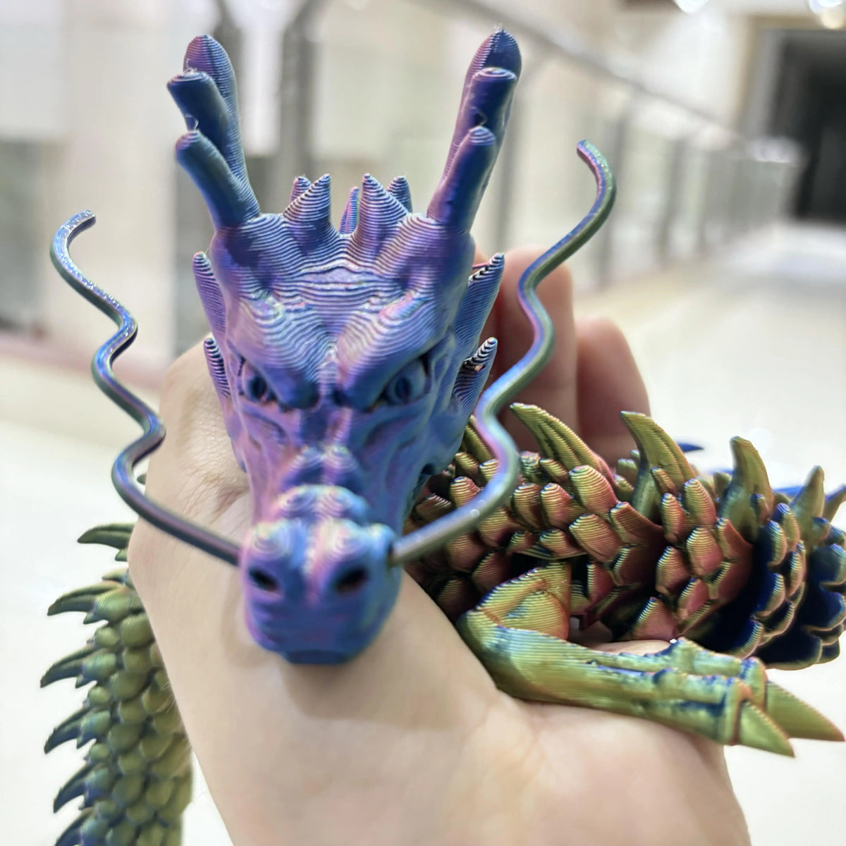 A hand holds a purple articulated 3D printed dragon figurine with curved horns and a textured green scale piece.