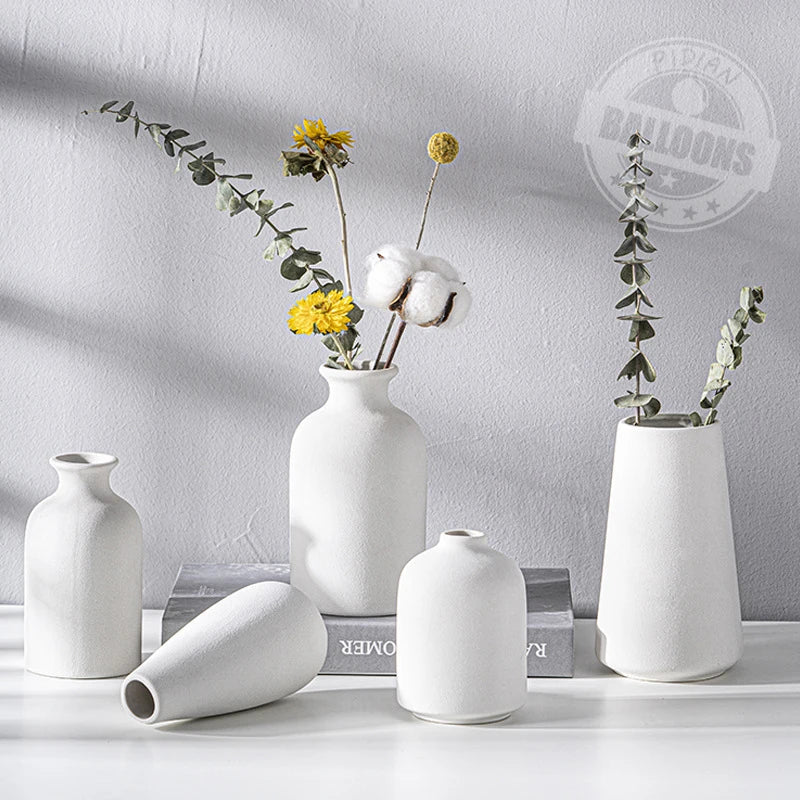 A white ceramic tabletop vase holds dried greenery and a fluffy white bloom against a light gray background.