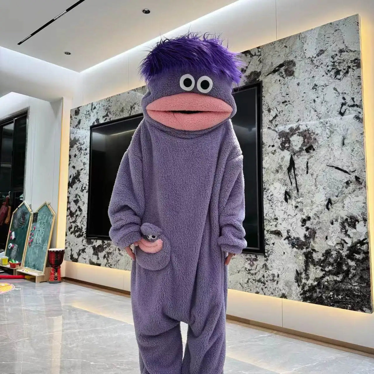 A purple, fuzzy monster coral fleece hooded sleepsuit with large eyes and a pink mouth standing in a modern room.