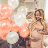 A pregnant woman smiles while holding a pink balloon garland in a bright indoor setting.
