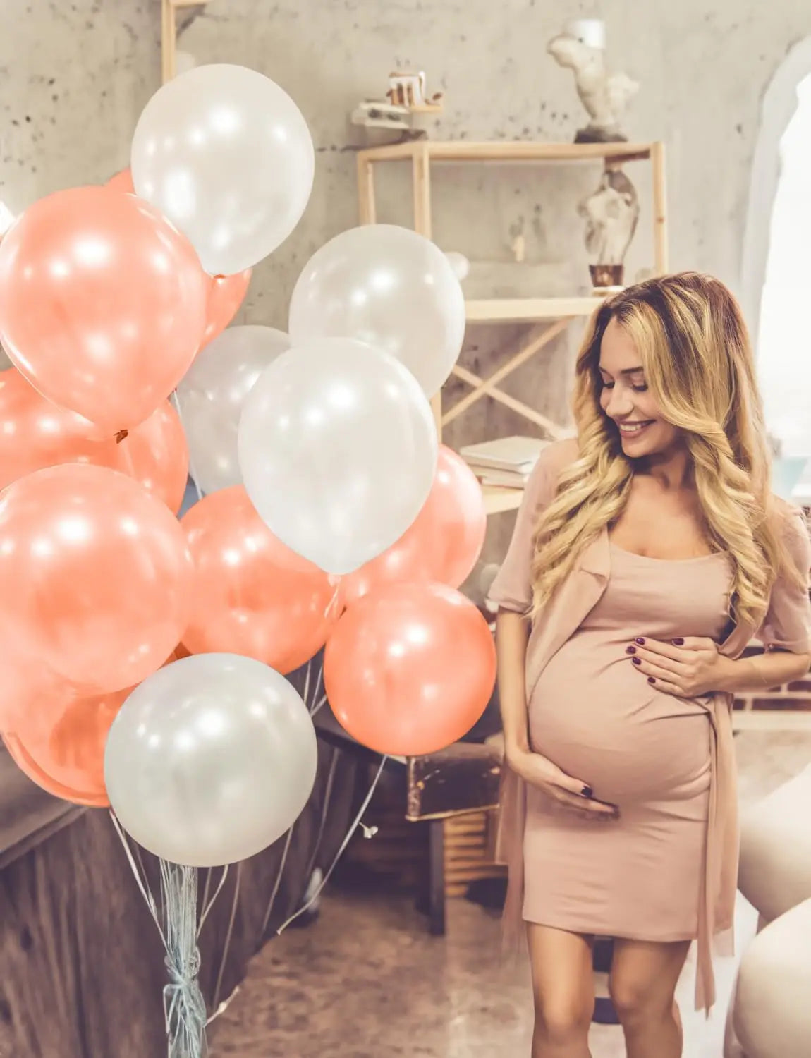 A pregnant woman smiles while holding a pink balloon garland in a bright indoor setting.