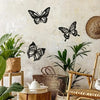 A set of three black metal butterfly wall art pieces displayed against a textured white wall with green plants and natural wo
