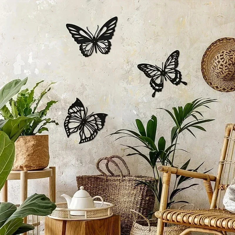 A set of three black metal butterfly wall art pieces displayed against a textured white wall with green plants and natural wo
