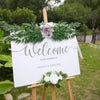 Un letrero blanco con caligrafía elegante da la bienvenida a la boda de un príncipe y princesa rodeados de ruscus italiano ve