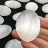 A hand holds a translucent white selenite palm crystal surrounded by other smooth, oval selenite pieces.