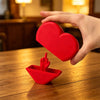 A hand holds a red heart-shaped novelty desk decor item, with a smaller red hand gesture piece resting on a wooden surface.