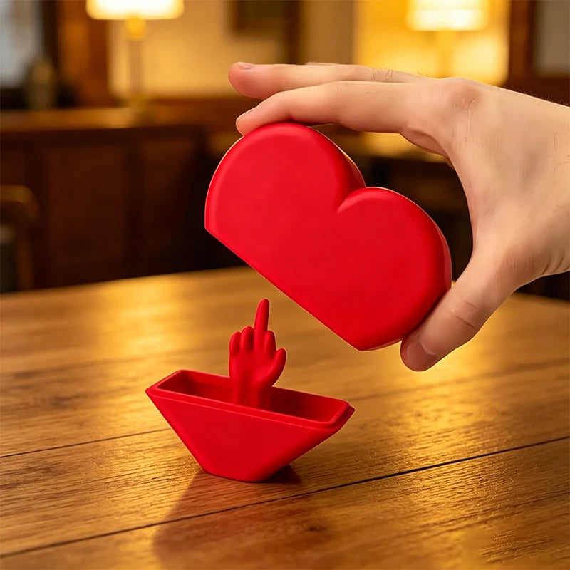 A hand holds a red heart-shaped novelty desk decor item, with a smaller red hand gesture piece resting on a wooden surface.