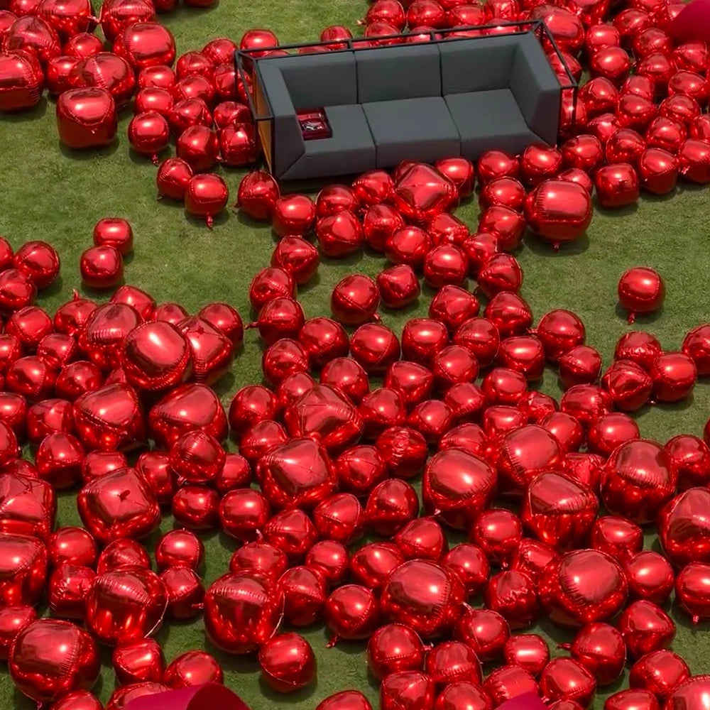 A large quantity of glossy red round foil balloons scattered on green grass with a dark gray sofa in the background.