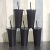 Four black reusable tumblers with lids and straws standing upright against a light wooden background.