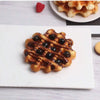 A golden waffle with blueberries is displayed on a white square plate, next to a plate of waffles and a raspberry.