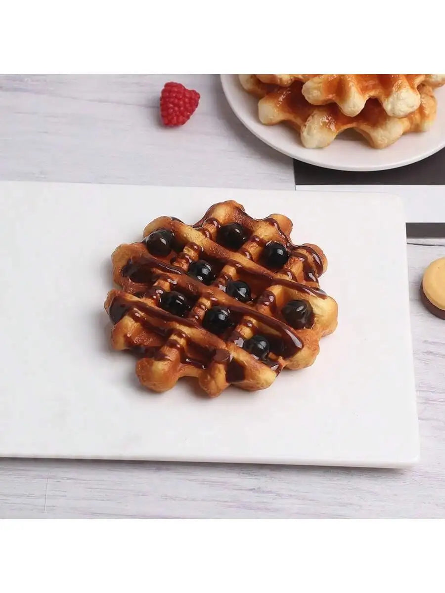 A golden waffle with blueberries is displayed on a white square plate, next to a plate of waffles and a raspberry.