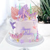 A pink mermaid birthday cake decorated with iridescent toppers shaped like fish tails, shells, and seaweed.