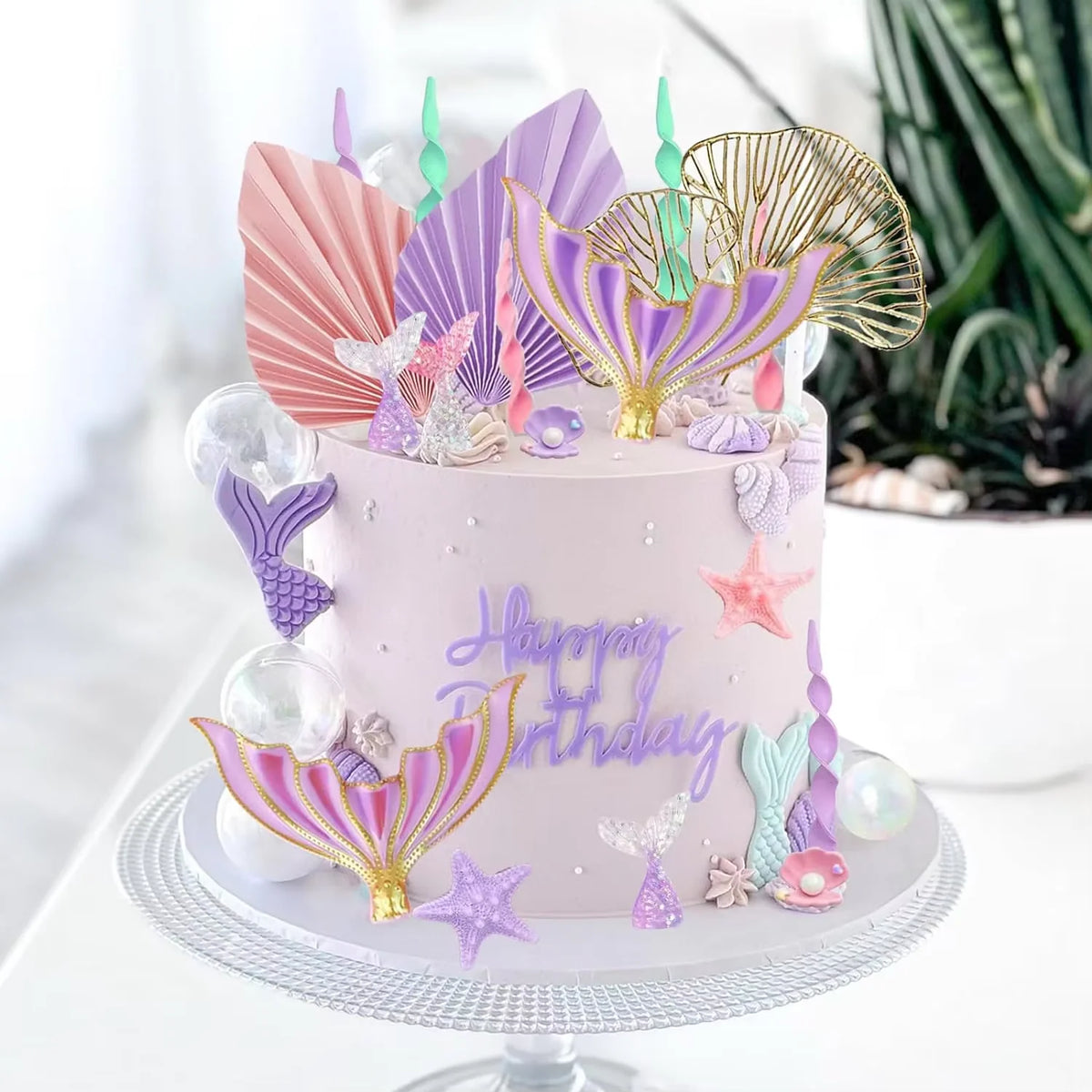 A pink mermaid birthday cake decorated with iridescent toppers shaped like fish tails, shells, and seaweed.