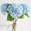 A hand holds a cluster of light blue hydrangea silk flower branches.