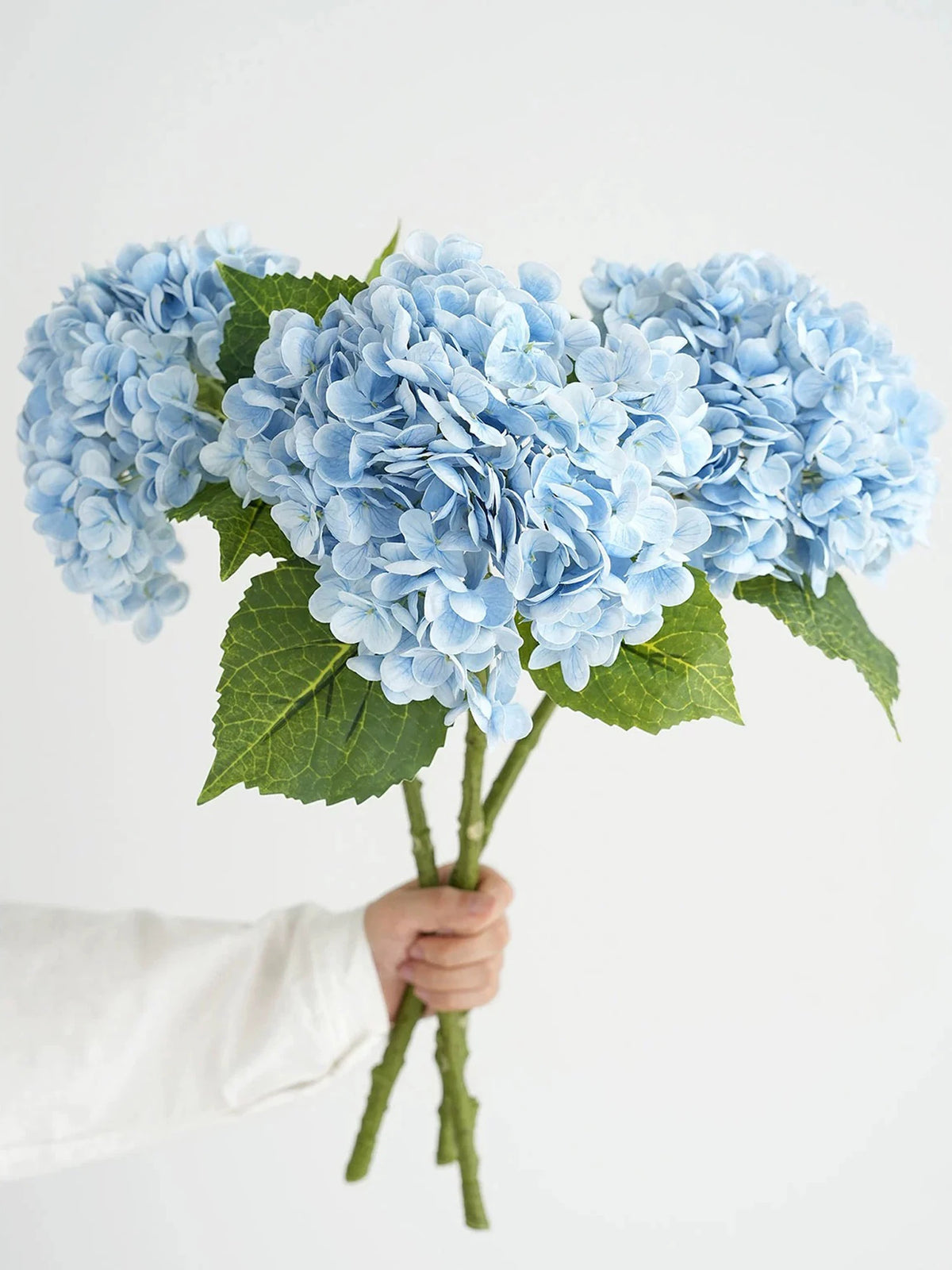 A hand holds a cluster of light blue hydrangea silk flower branches.