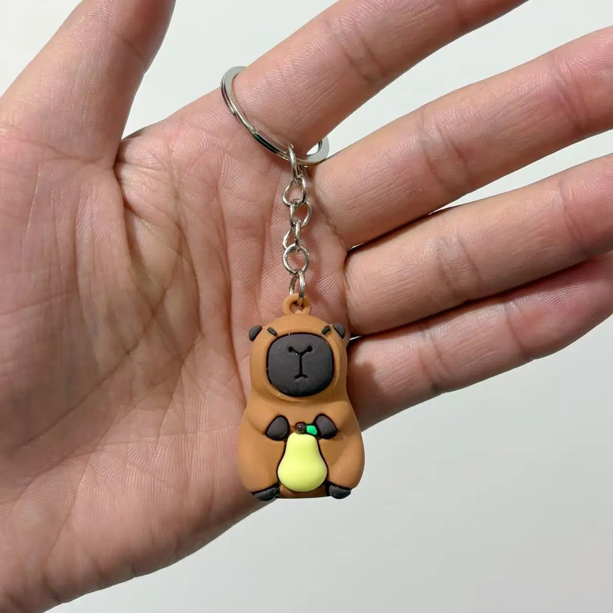 Un petit pendentif de capybara brun rond avec des accents jaunes est tenu par les doigts d'une personne.