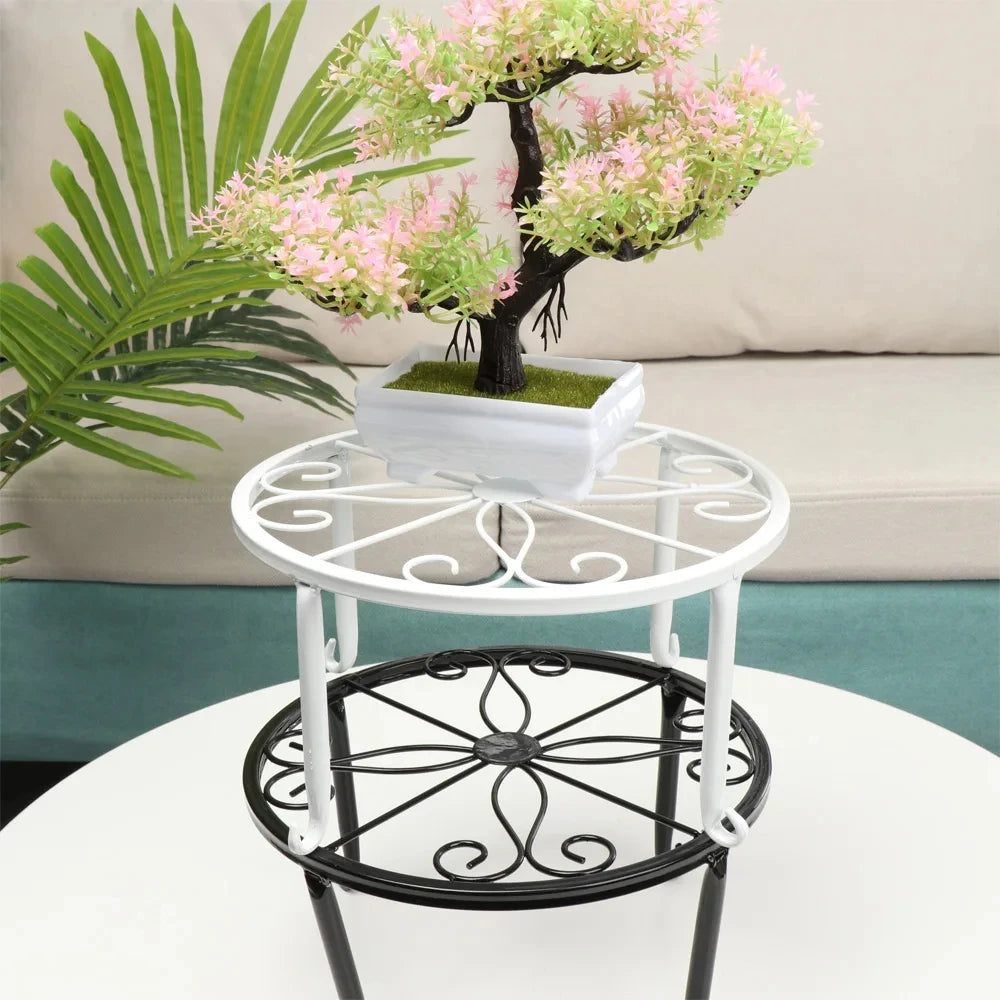 A white decorative metal plant stand with a black saucer holding a small tree and pink blossoms.