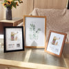 A set of five rustic wooden photo frames displayed on a light wooden table with greenery and soft textiles in the background.