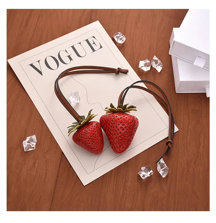 A brown leather strap charm featuring a 3D strawberry design and clear ice crystals, displayed on a paper labeled VOGUE.