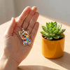 A hand holds a sparkling Yorkshire Terrier acrylic pendant necklace next to a potted succulent plant.
