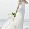 A woman in a white dress holds an artificial delphinium bouquet near the ocean.