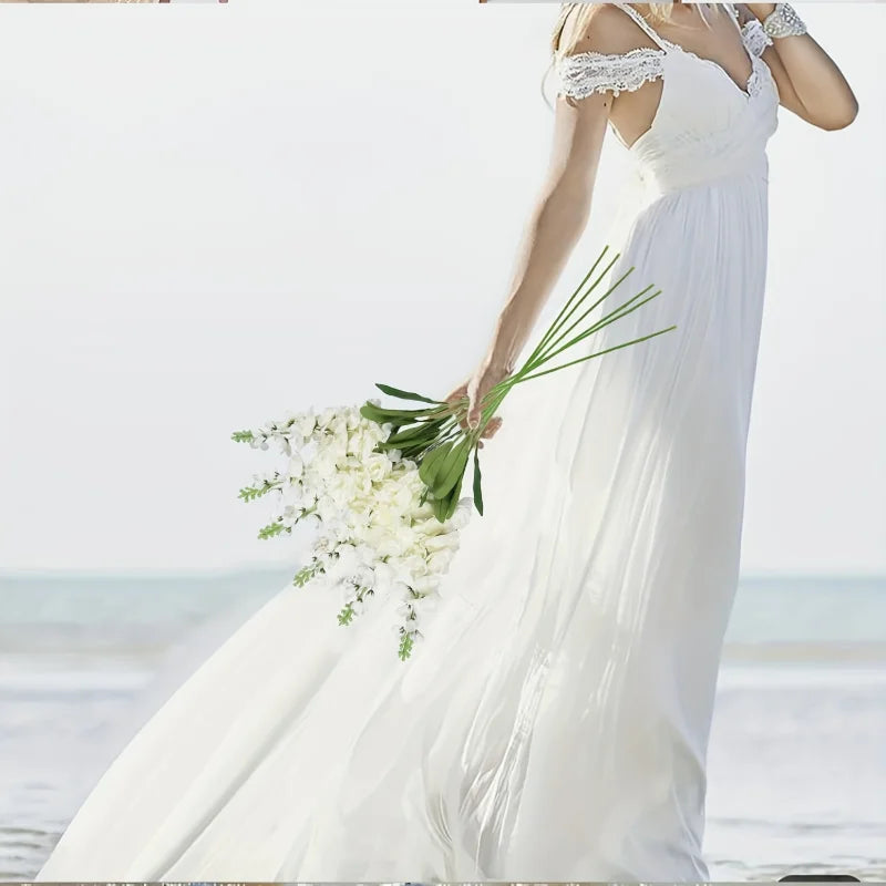 A woman in a white dress holds an artificial delphinium bouquet near the ocean.