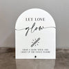 A white acrylic sign with black script that reads Let Love glow and features an illustration of a glow stick.