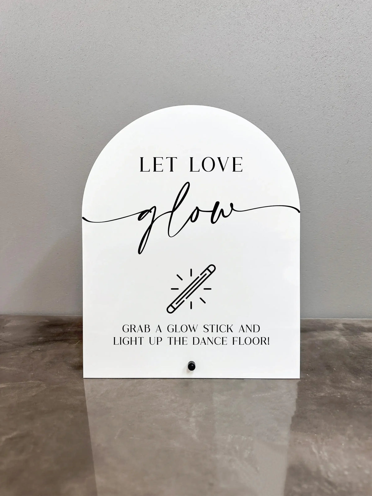 A white acrylic sign with black script that reads Let Love glow and features an illustration of a glow stick.