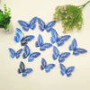 A collection of twelve blue butterfly wall stickers scattered on a white surface next to a potted plant.
