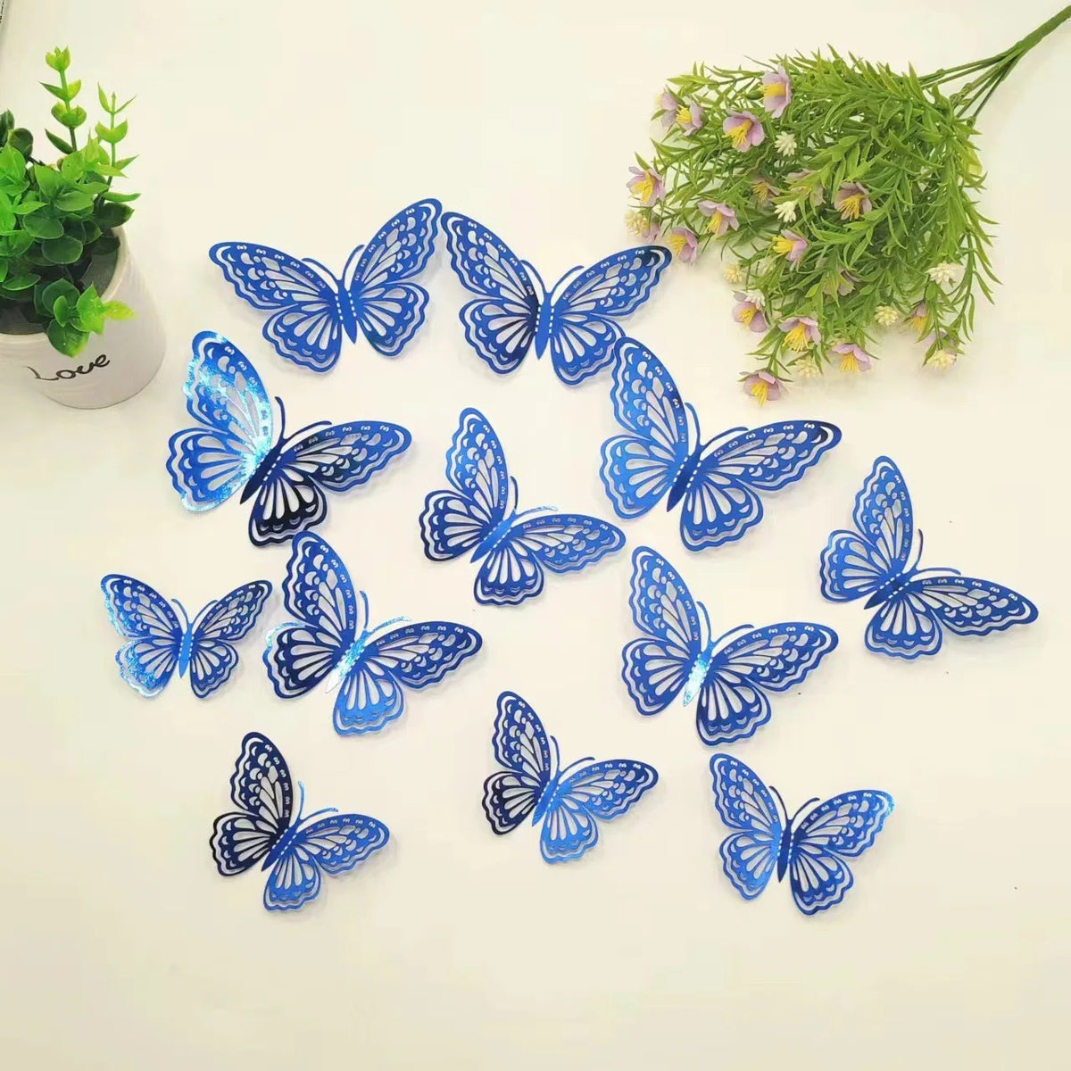 A collection of twelve blue butterfly wall stickers scattered on a white surface next to a potted plant.