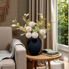 A dark blue vase filled with light pink and green floral arrangements sits on a wooden table next to a beige armchair.