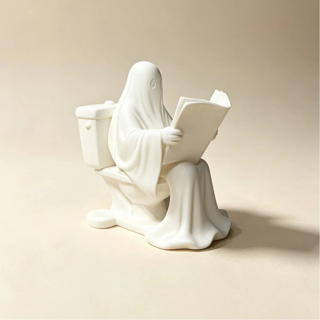 A white statue of a ghost reading a book sitting on a toilet.