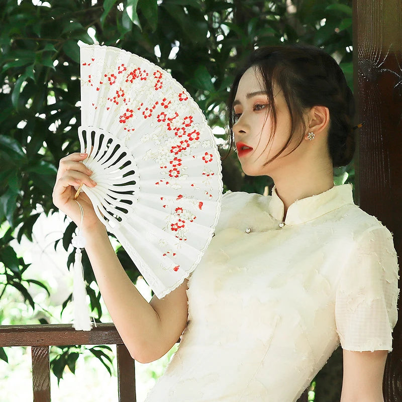 A woman in a white top holds an elegant plum blossom folding fan outdoors.