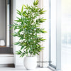 An artificial bamboo branch with green leaves in a white pot placed next to a window.