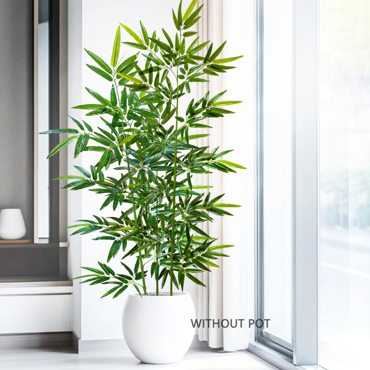An artificial bamboo branch with green leaves in a white pot placed next to a window.