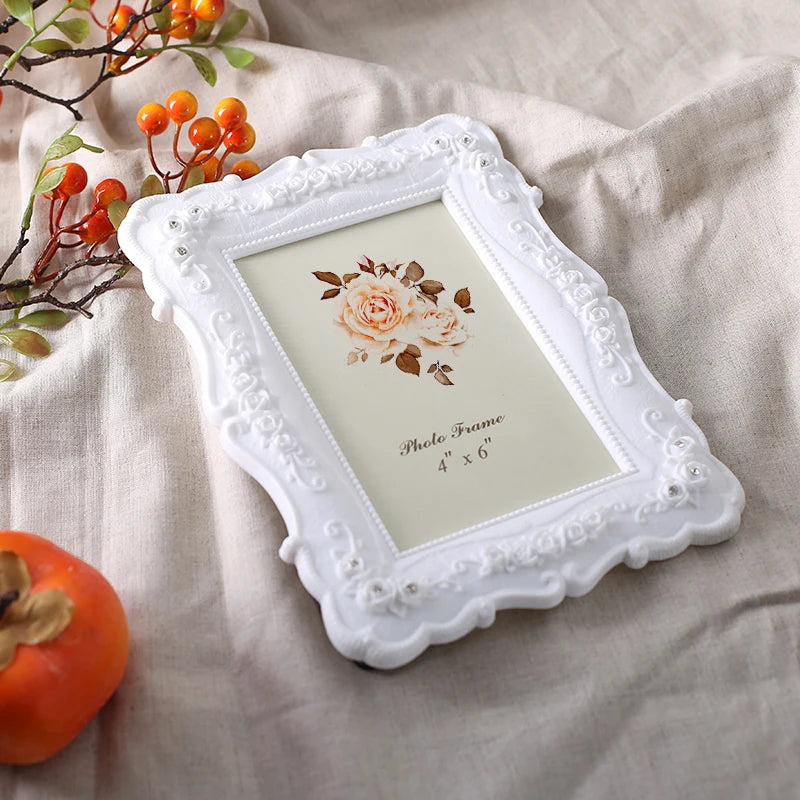 A white relief photo frame with a decorative border and a floral design is displayed on a beige fabric next to an orange frui