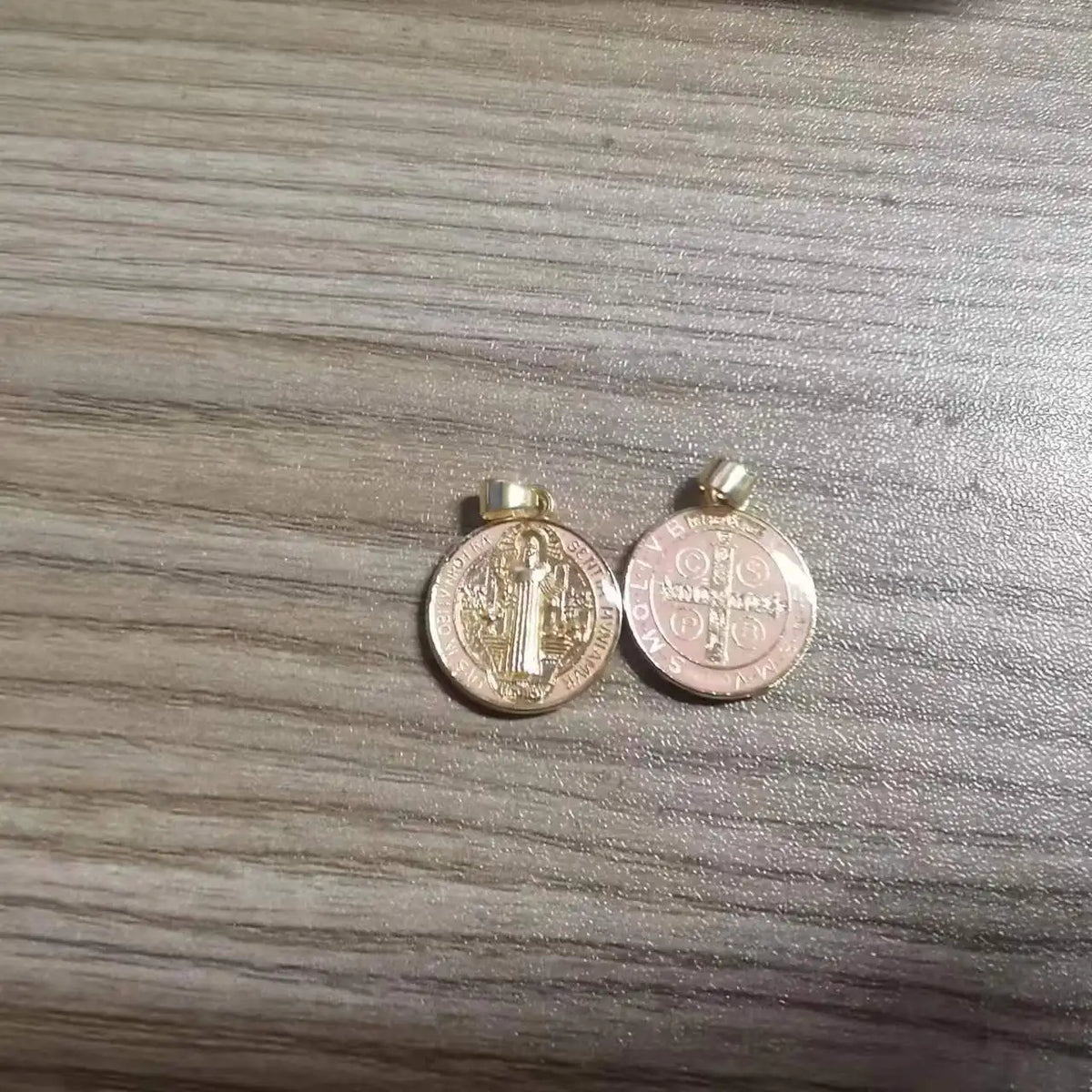A pair of gold-colored St. Benedict religious pendants or medals resting on a wooden surface.