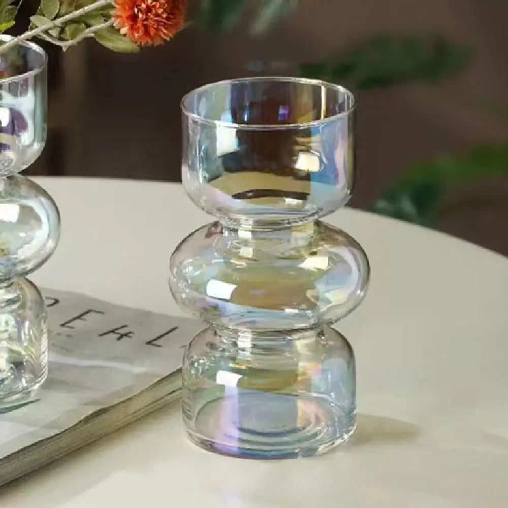 A clear glass flower vase with stacked, rounded sections sits on a white surface next to a book and greenery.