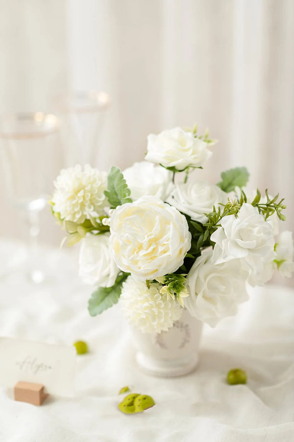 A bouquet of white artificial dahlia flowers and green foliage in a white vase on a white cloth.