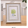 A framed watercolor painting of green foliage in a blue vase displayed on a wooden table.