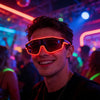A young man wearing neon orange light up EL wire party glasses smiles in a dimly lit party environment.